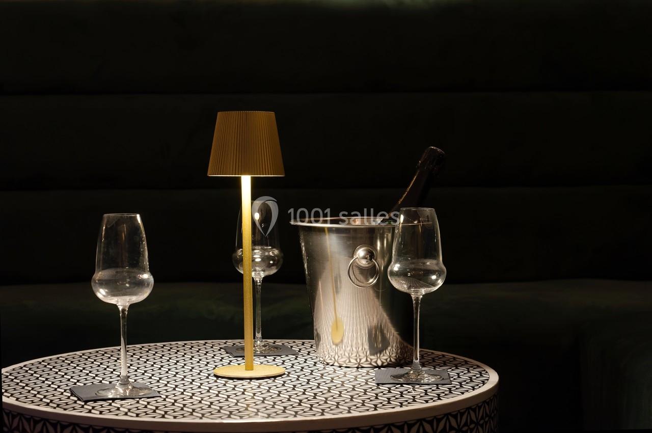 Table avec une lampe allumée, deux verres à pied vides et un seau contenant une bouteille de champagne.