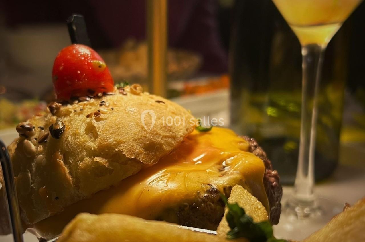 Burger garni de fromage fondu et d'une tomate cerise, accompagné de frites dorées et d'un cocktail en arrière-plan.