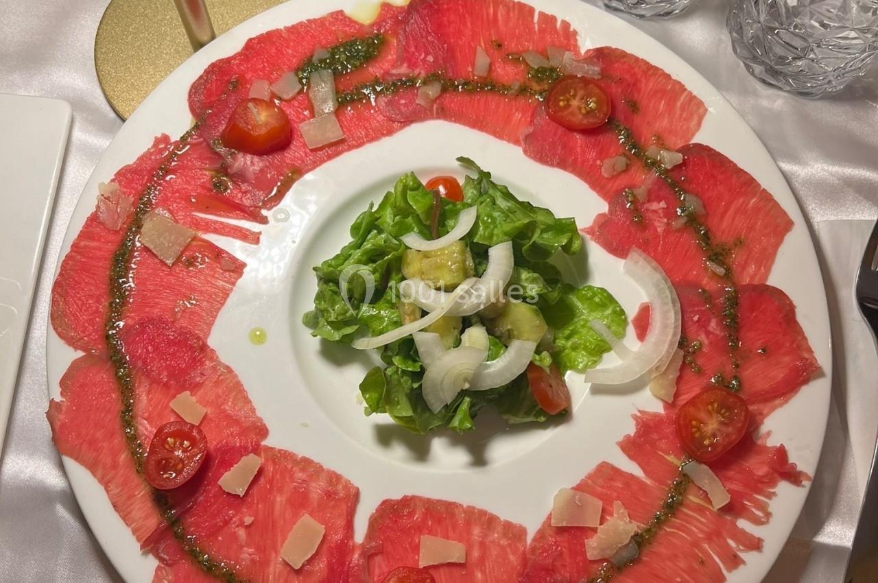 Assiette de carpaccio de bœuf entourant une salade verte garnie de tomates cerises, copeaux de parmesan et sauce.