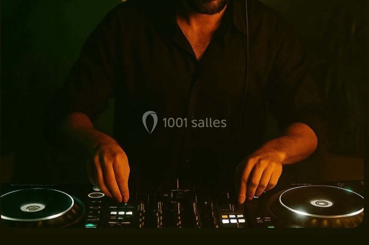 Un DJ en chemise noire mixe de la musique sur une table de mixage éclairée dans une ambiance sombre.
