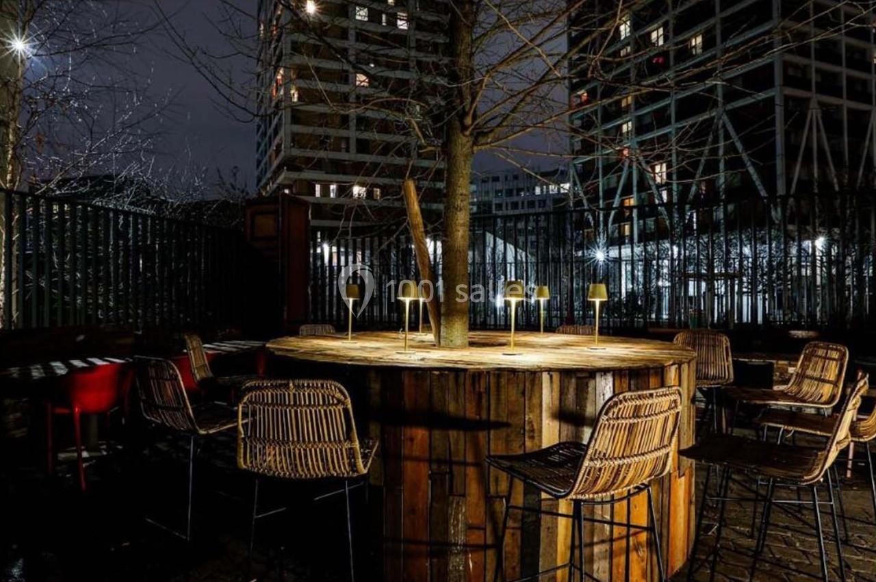 Terrasse extérieure nocturne avec table ronde en bois entourée de chaises en rotin, éclairée par des lampes.
