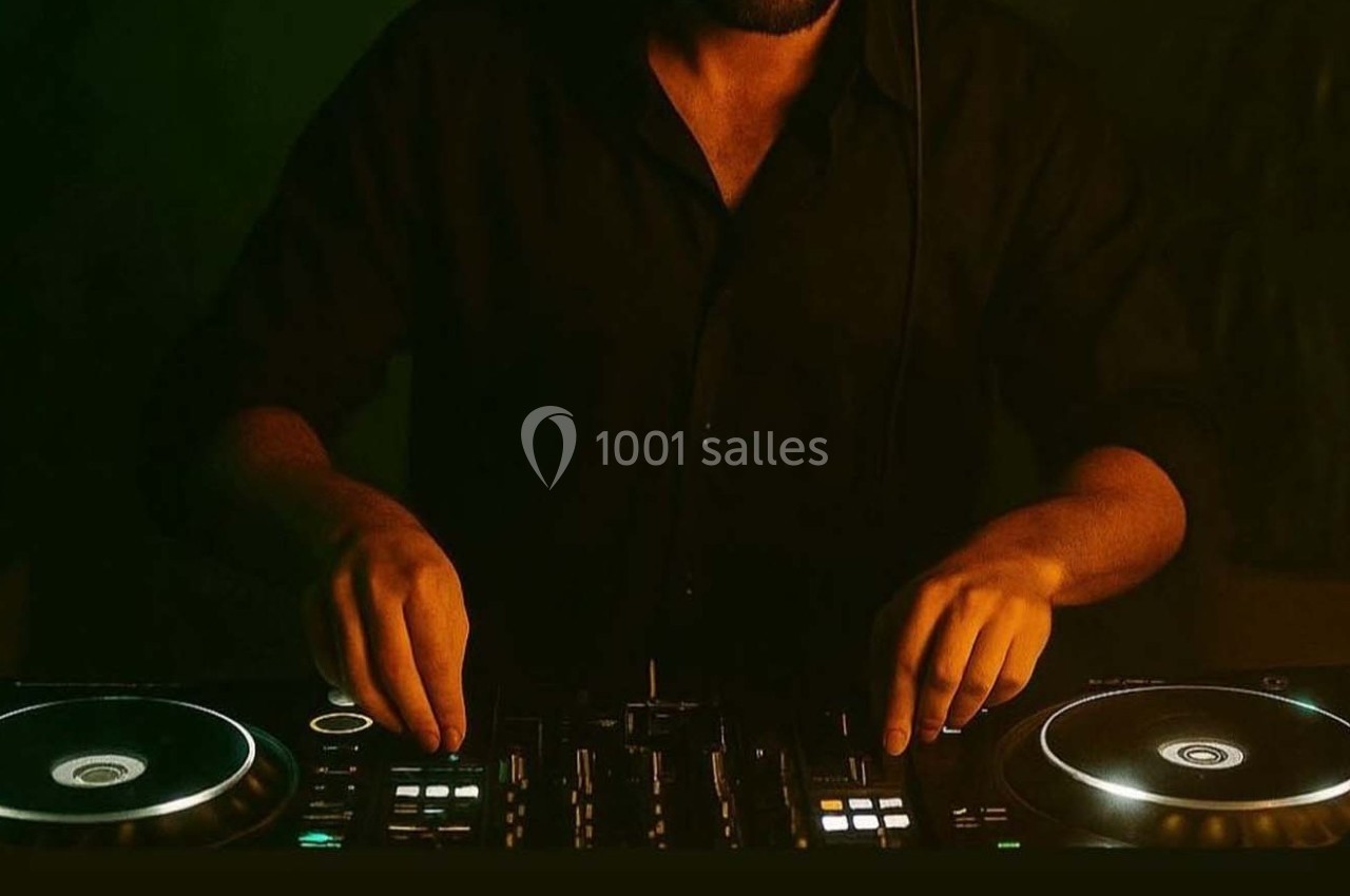 Un DJ en chemise noire manipule une table de mixage éclairée dans une ambiance sombre.