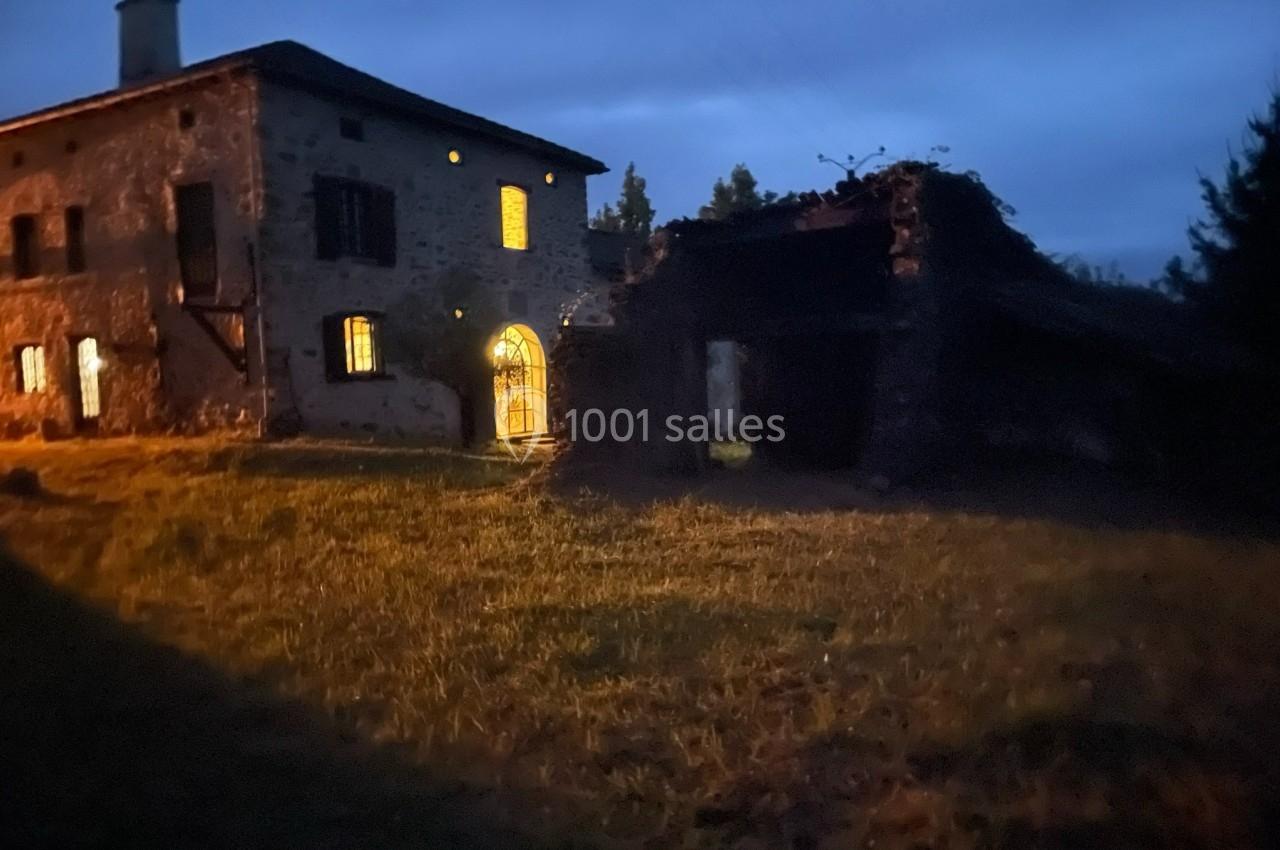 Maison en pierre éclairée à la tombée de la nuit, avec une annexe en ruine et un jardin herbeux au premier plan.