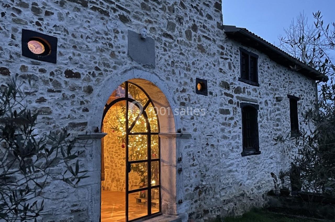 Façade en pierre d'une maison éclairée à la tombée de la nuit, avec une porte vitrée laissant entrevoir un intérieur…