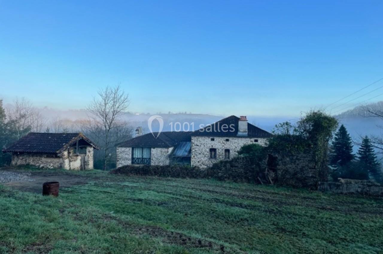 Maison en pierre entourée de verdure avec un ciel bleu dégagé et un paysage légèrement brumeux en arrière-plan.