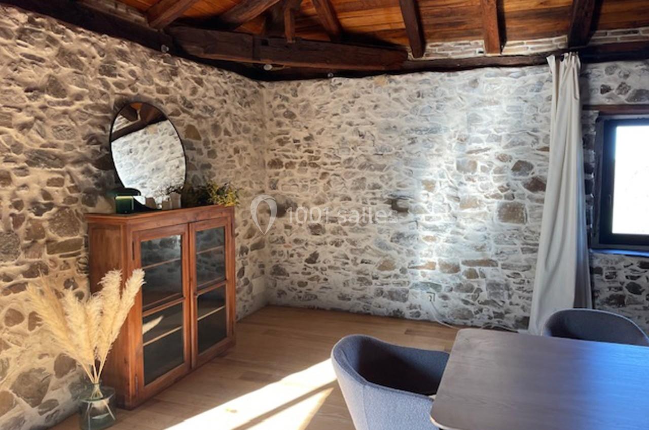 Mur en pierre apparente avec meuble en bois, miroir rond, chaise et table, éclairés par une fenêtre à droite.