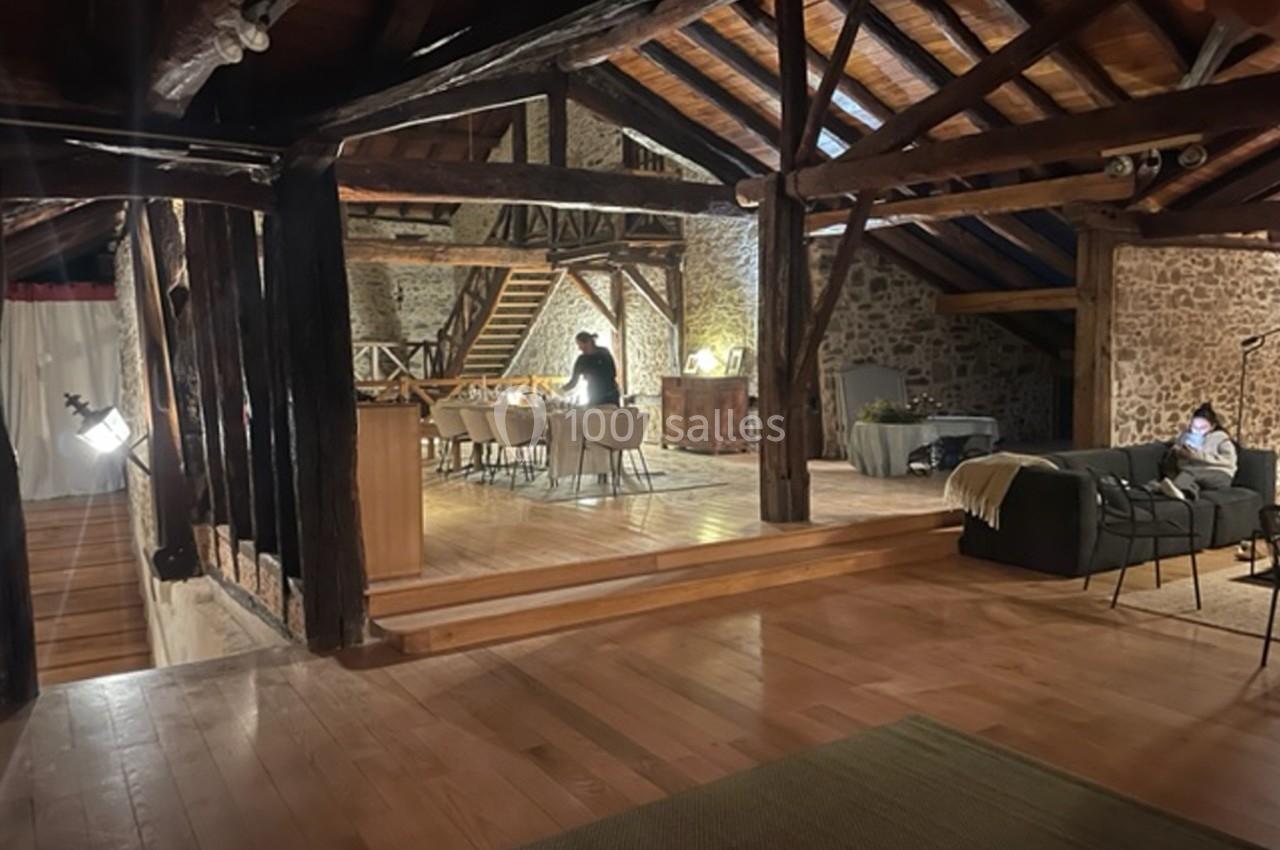 Vue d'un intérieur rustique avec poutres apparentes, espace repas éclairé et coin salon dans une ambiance chaleureuse.