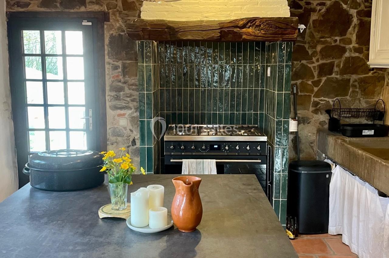 Cuisine rustique avec murs en pierre, cuisinière à gaz, plan de travail en béton et décoration simple avec fleurs et bougies.