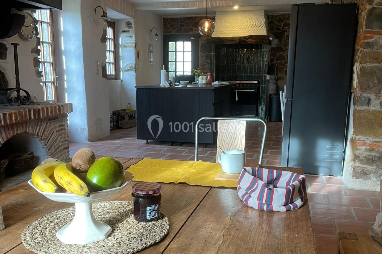 Cuisine rustique avec murs en pierre, table en bois garnie de fruits, pot et nappe, et îlot central moderne en arrière-plan.