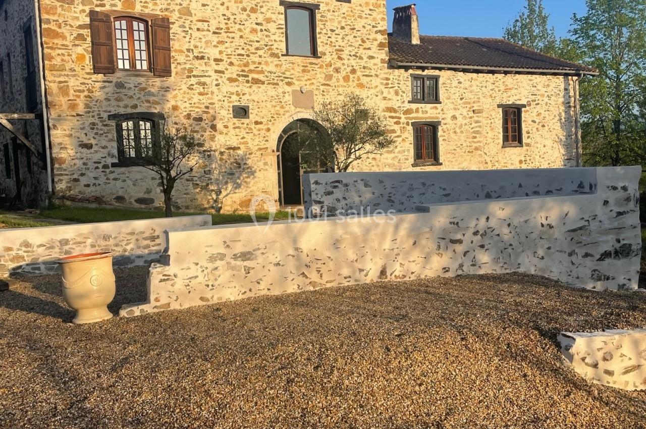 Maison en pierre avec volets en bois, entourée d'une cour gravillonnée et d'un mur bas, sous un ciel dégagé.
