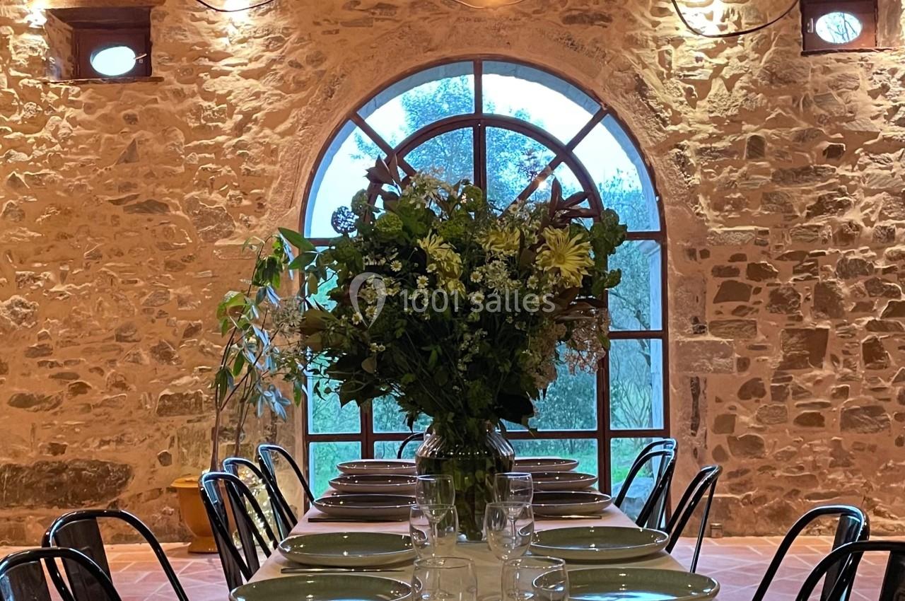 Grande table dressée avec vaisselle et bouquet de fleurs, dans une salle en pierre avec une grande fenêtre en arche.