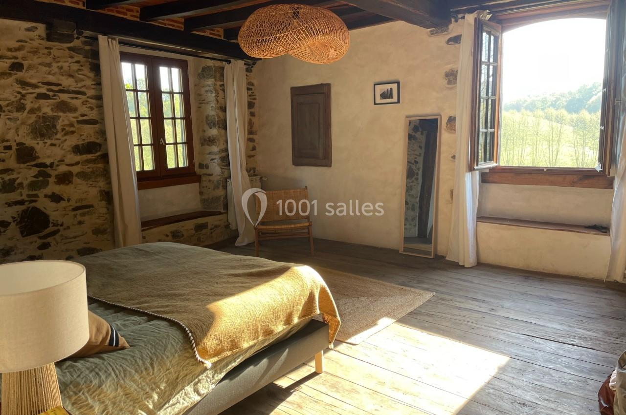 Chambre lumineuse avec murs en pierre, parquet en bois, lit, fauteuil en osier, fenêtres ouvertes sur un paysage verdoyant.