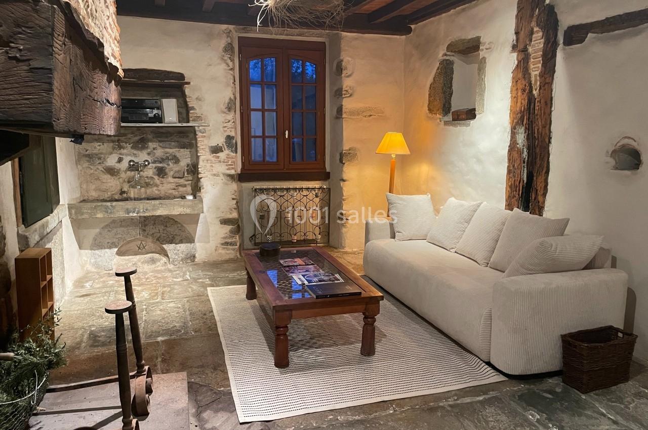 Salon rustique avec murs en pierre, poutres apparentes, canapé blanc, table basse en bois et lampe allumée.