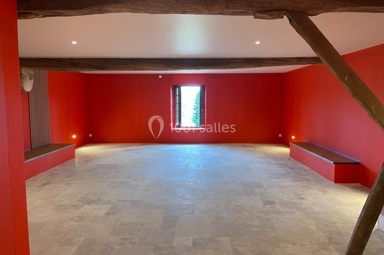 Salle lumineuse avec murs rouges, poutres apparentes en bois, sol en pierre claire et une fenêtre centrée.