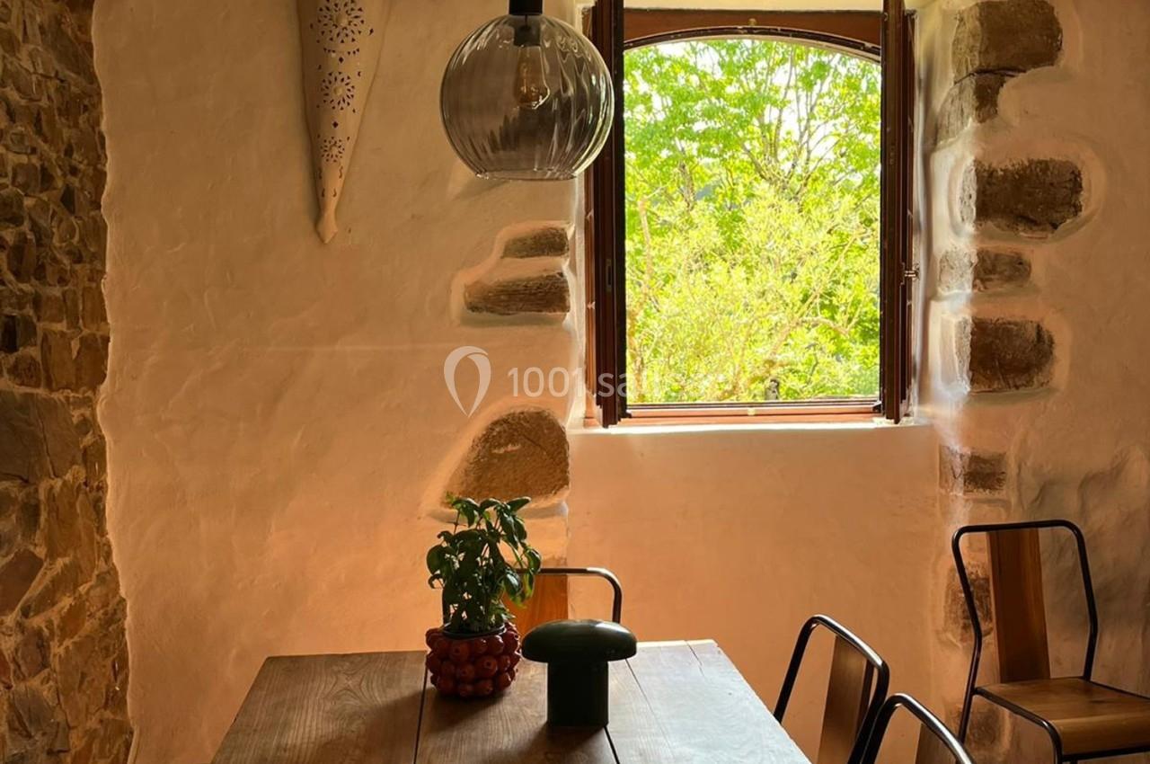 Salle à manger rustique avec table en bois, chaises métalliques, plante en pot et fenêtre ouverte sur un jardin verdoyant.