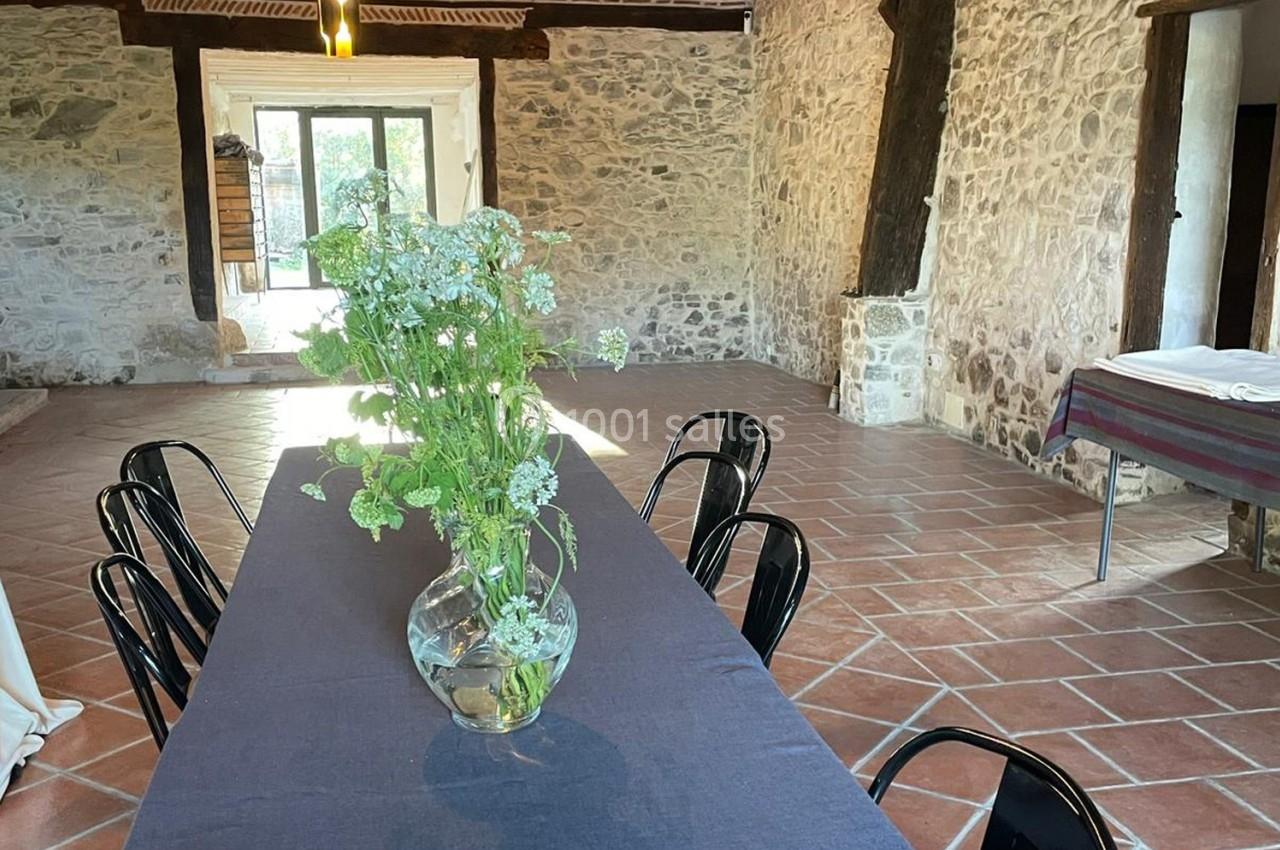 Salle en pierre avec une table noire, des chaises en métal et un vase de fleurs blanches au centre.