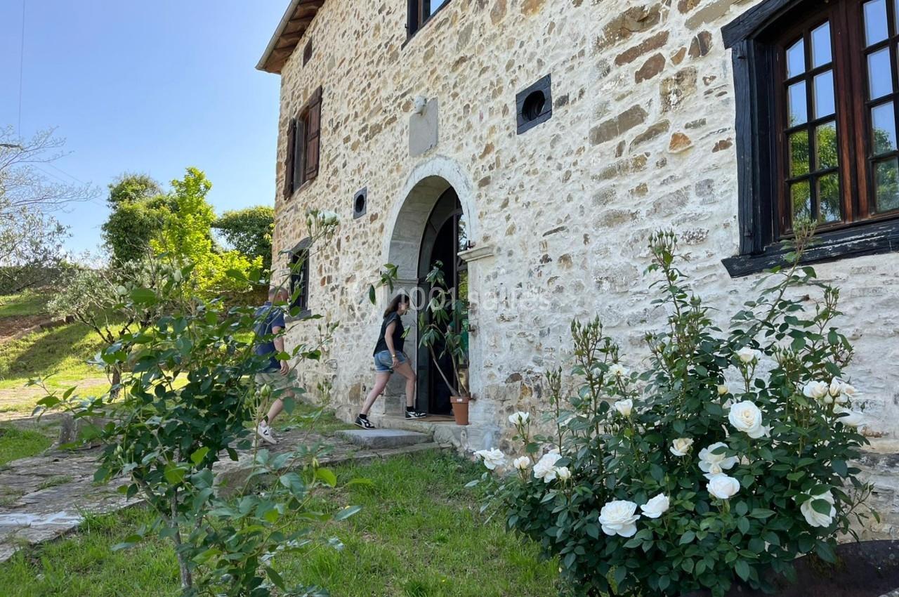 Façade en pierre d'une maison avec des rosiers blancs au premier plan et deux personnes entrant par la porte.