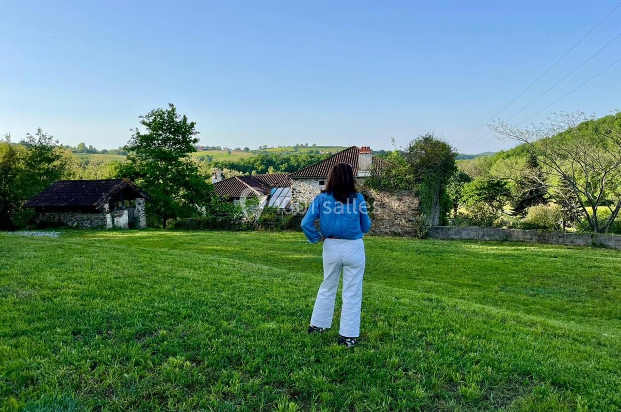 Une femme de dos, debout sur une pelouse, regarde un paysage rural avec des arbres et des bâtiments en arrière-plan.