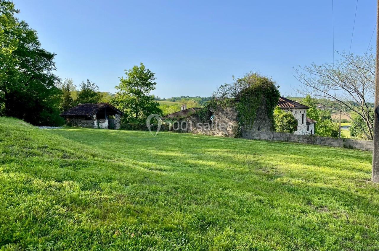 Terrain herbeux avec des arbres, un mur en pierre, des bâtiments anciens et un paysage rural en arrière-plan.