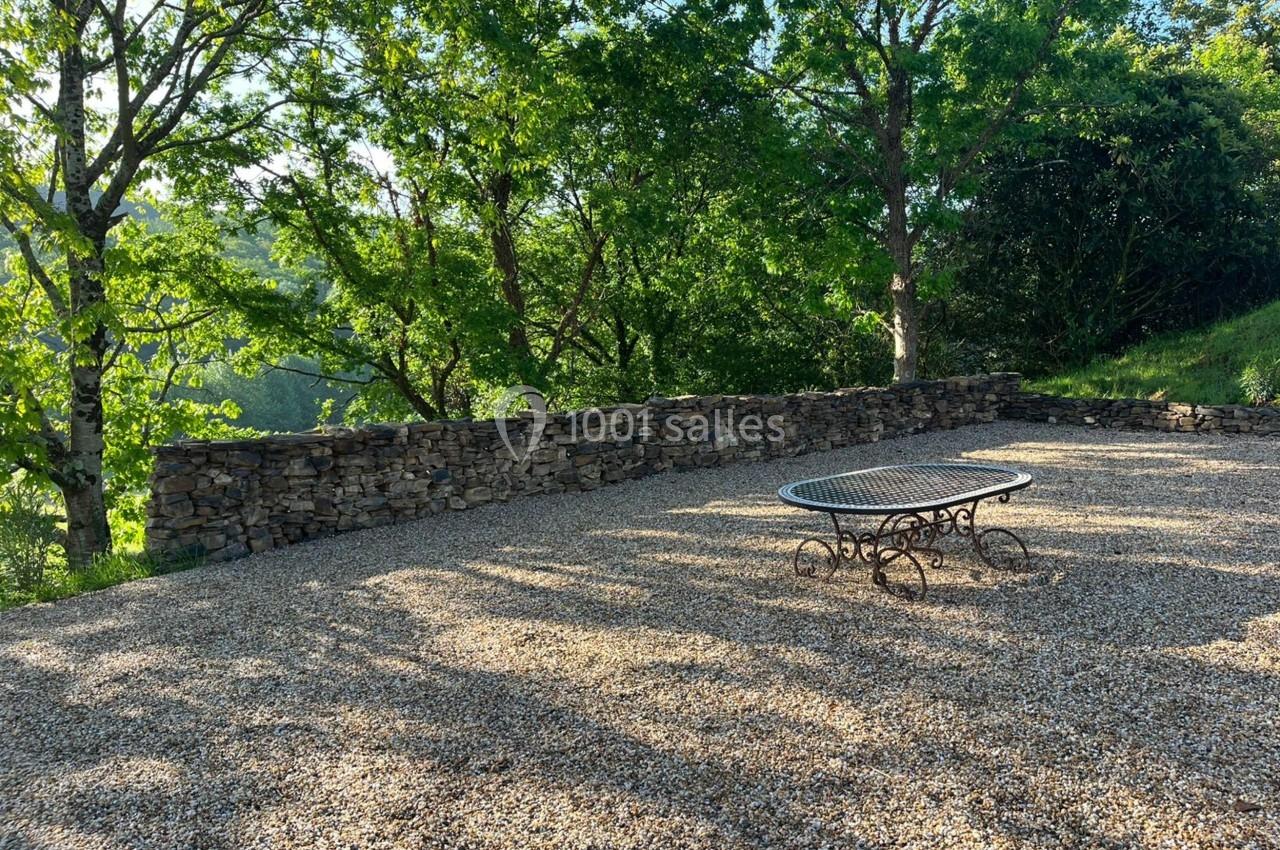 Terrasse en gravier avec une table en fer forgé, entourée d'arbres et d'un muret en pierre sèche.