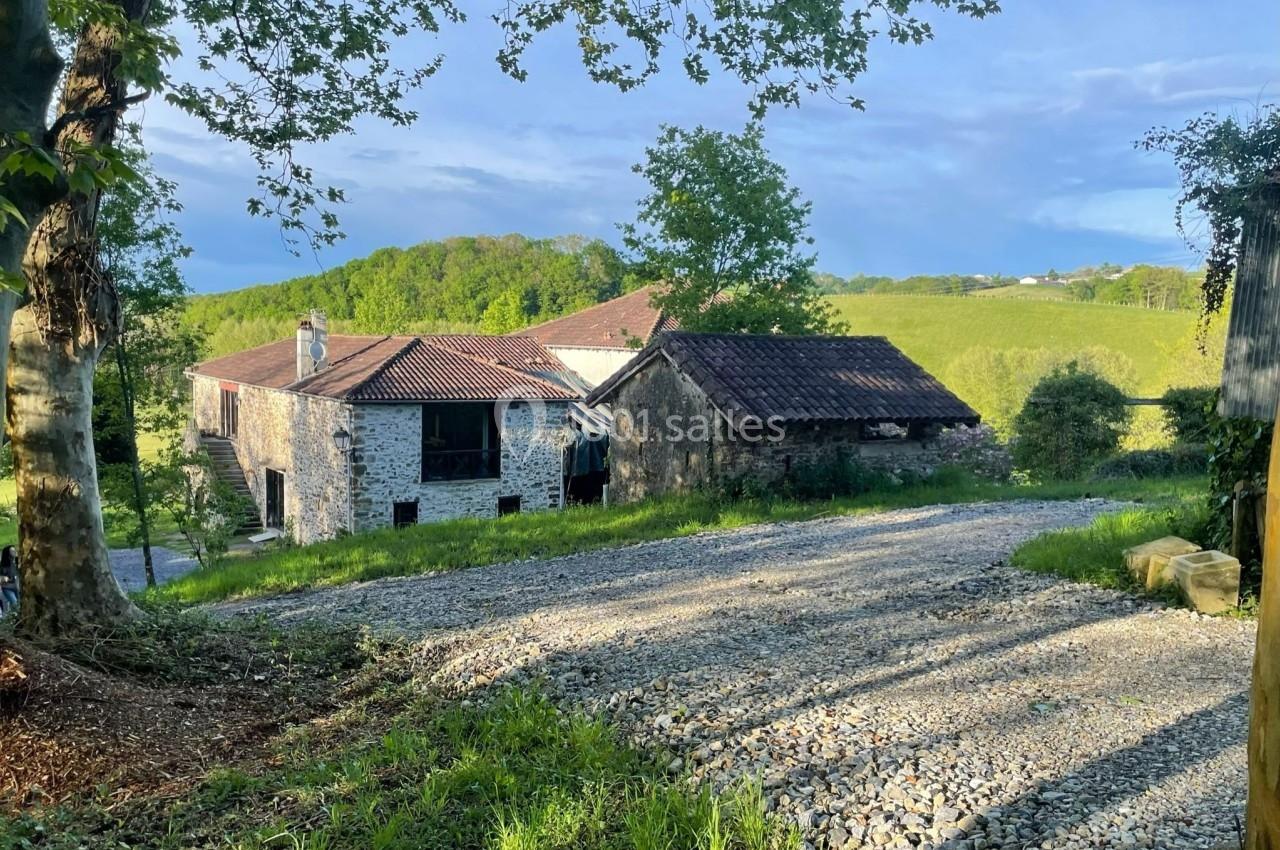 Chemin de gravier menant à des bâtiments en pierre entourés de verdure et de collines sous un ciel dégagé.