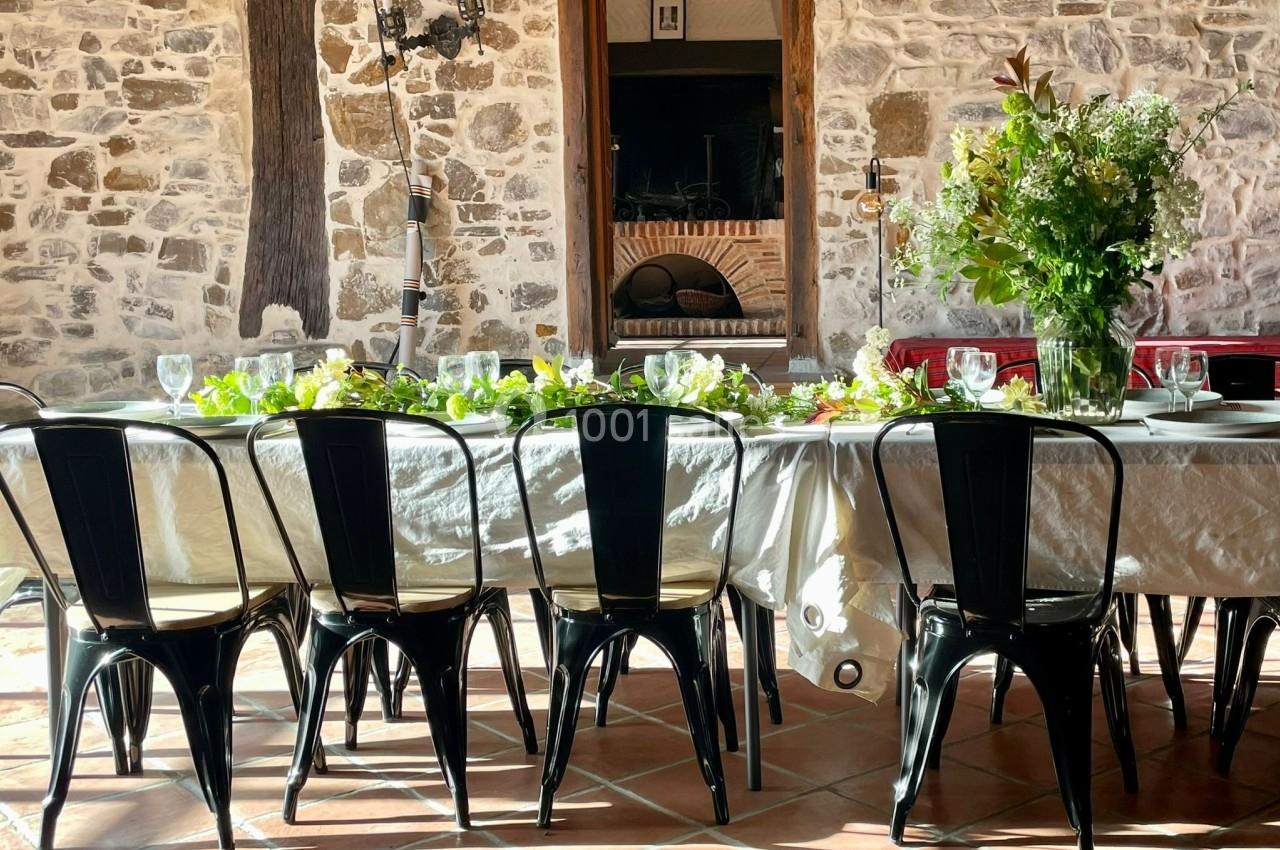 Table dressée avec une nappe blanche, chaises noires et décoration florale, dans une pièce aux murs en pierre.