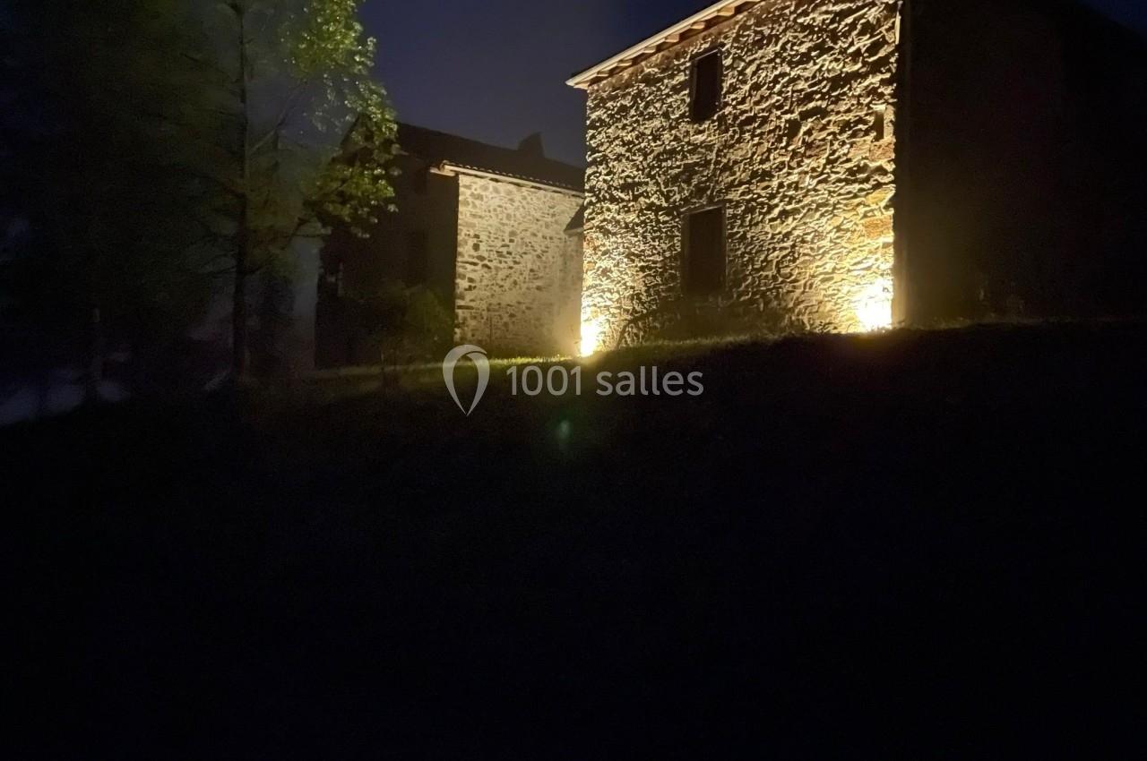 Façade en pierre d'une maison éclairée par des lumières au sol, entourée d'arbres, de nuit.