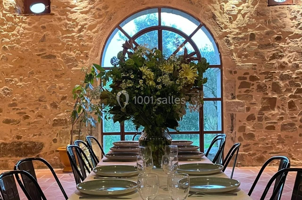 Grande table dressée avec vaisselle et verres, décorée d'un bouquet de fleurs, devant une fenêtre voûtée en pierre.