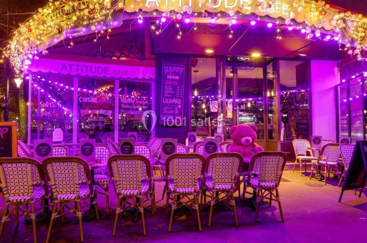Terrasse d’un café décorée de guirlandes lumineuses violettes, avec des chaises en osier et un grand ours en peluche.