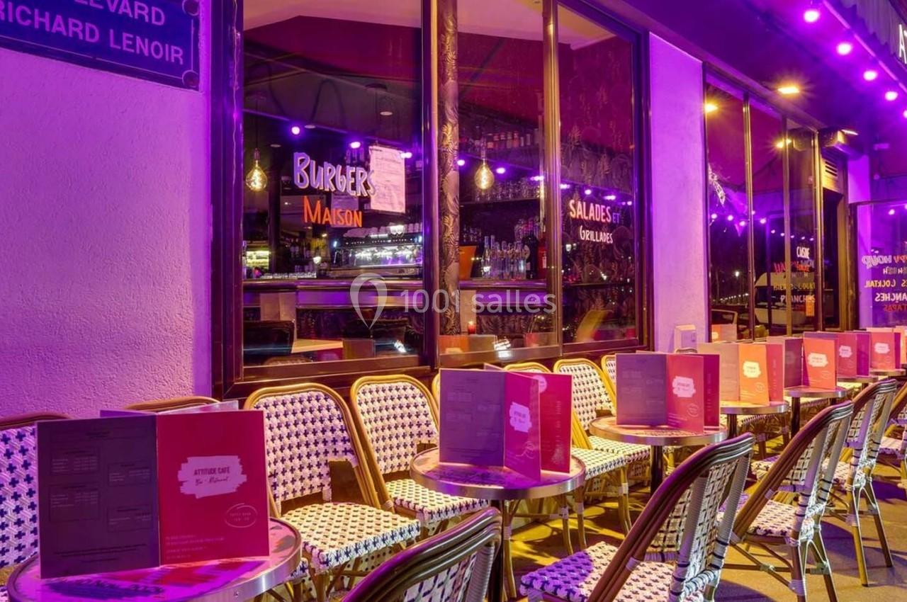 Terrasse de restaurant avec tables et chaises en osier, éclairée par des lumières violettes en soirée.
