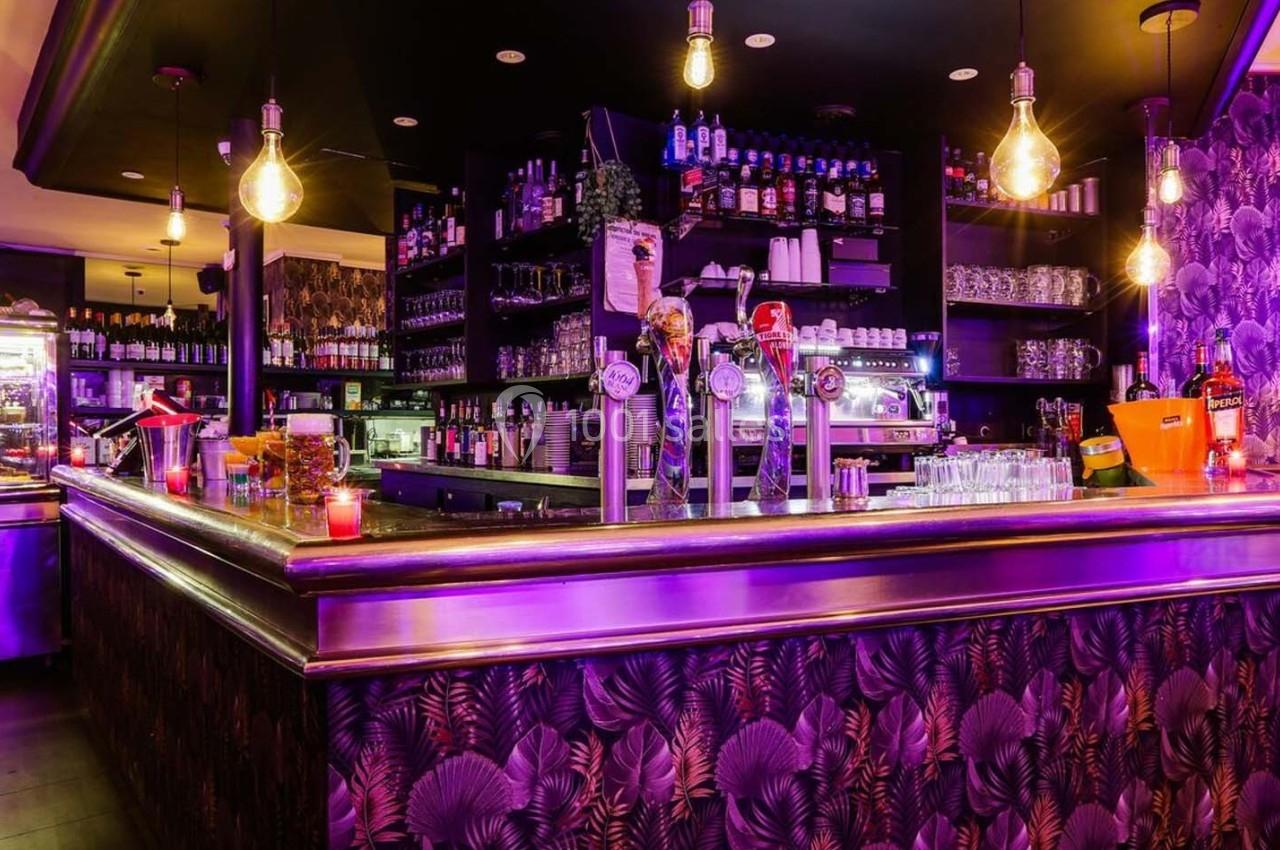 Bar avec comptoir éclairé, décorations murales à motifs, bouteilles alignées et lumières suspendues dans une ambiance…