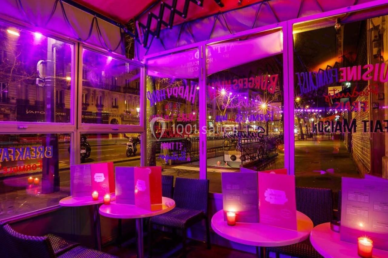 Terrasse d'un café-restaurant éclairée par une lumière violette, avec des tables, menus et bougies allumées.