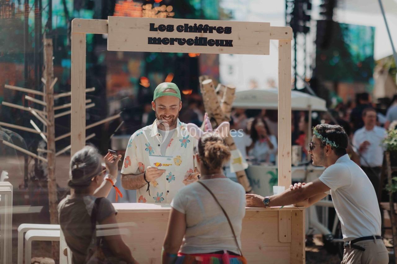 Un homme souriant derrière un stand en bois interagit avec trois personnes dans un environnement festif en extérieur.