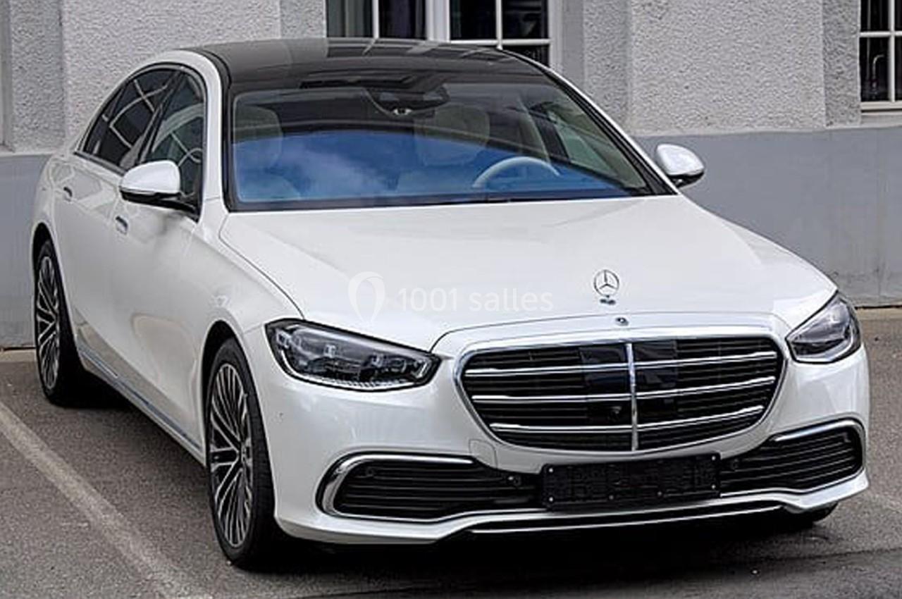 Berline blanche stationnée devant un bâtiment, vue de face, avec des détails de calandre et phares visibles.