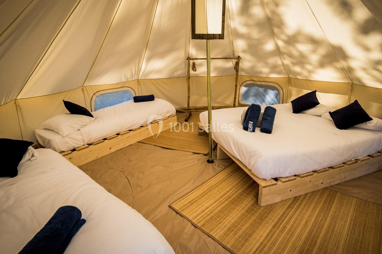 Intérieur d'une tente aménagée avec trois lits simples, draps blancs, coussins noirs et serviettes pliées.