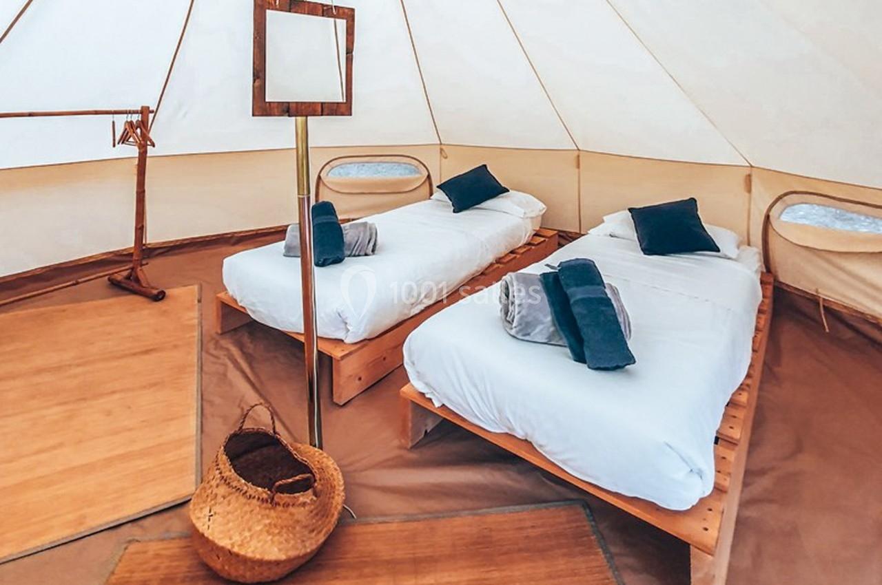 Intérieur d'une tente aménagée avec deux lits simples, draps blancs, coussins bleus et panier en osier au sol.