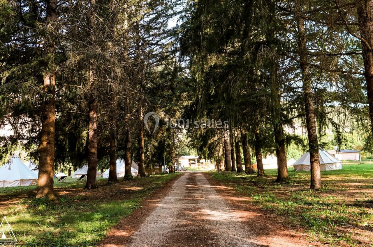 Chemin de terre bordé d'arbres dans une forêt, avec des tentes blanches visibles en arrière-plan.