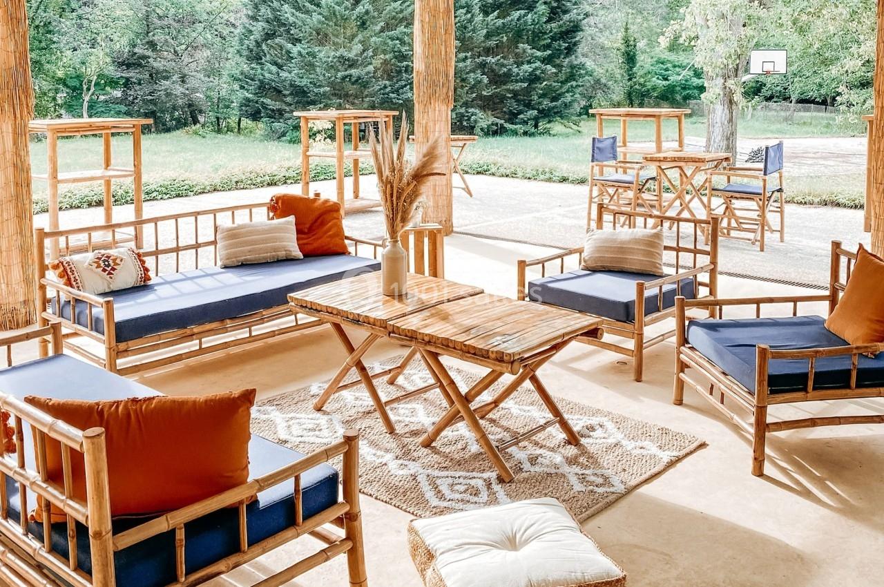 Salon extérieur en bambou avec coussins colorés, tapis beige et vue sur un jardin arboré avec un panier de basket.