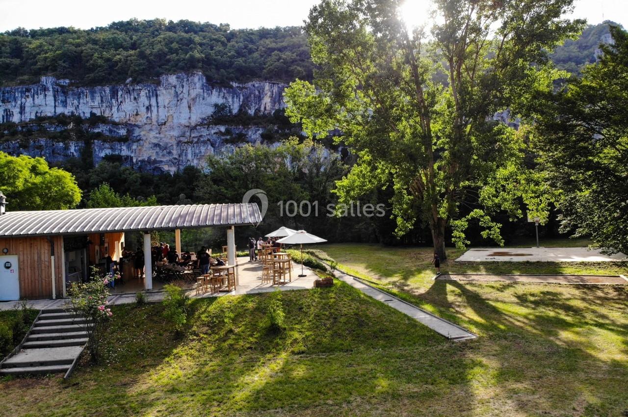 Terrasse d'un bâtiment en bois avec des tables, entourée de verdure, au pied d'une falaise ensoleillée.