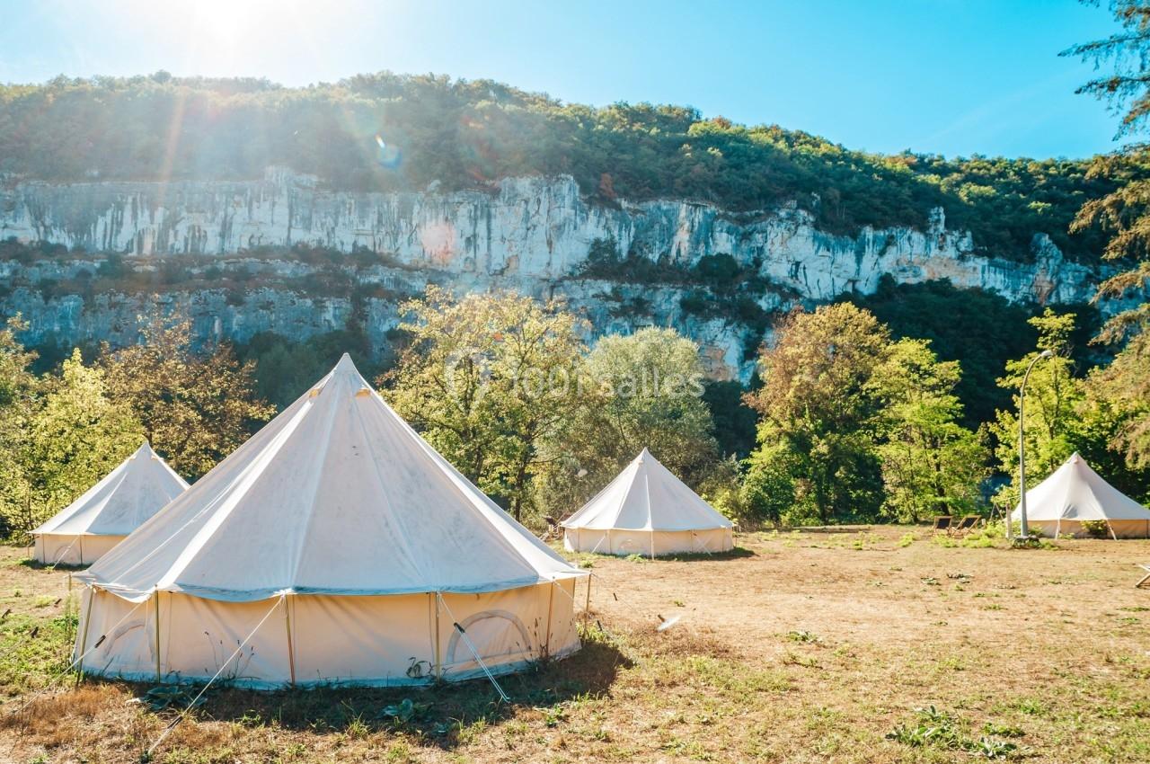 Tentes blanches installées sur un terrain herbeux, entourées d'arbres et de falaises sous un ciel ensoleillé.
