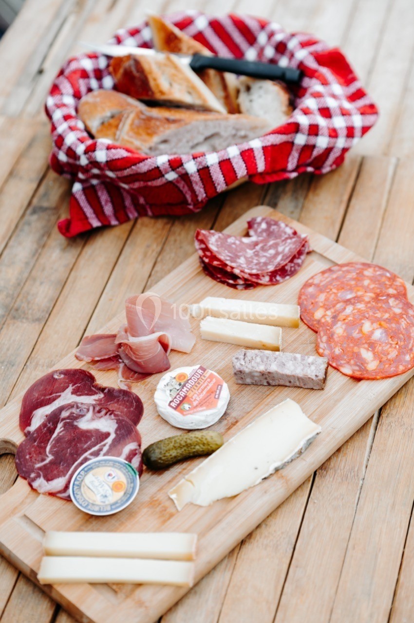 Plateau en bois avec charcuterie, fromages, cornichon et pain dans un panier en arrière-plan sur une table en bois.