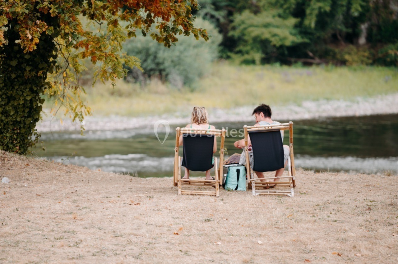 Deux personnes assises sur des chaises longues en bois au bord d'une rivière, entourées de végétation.