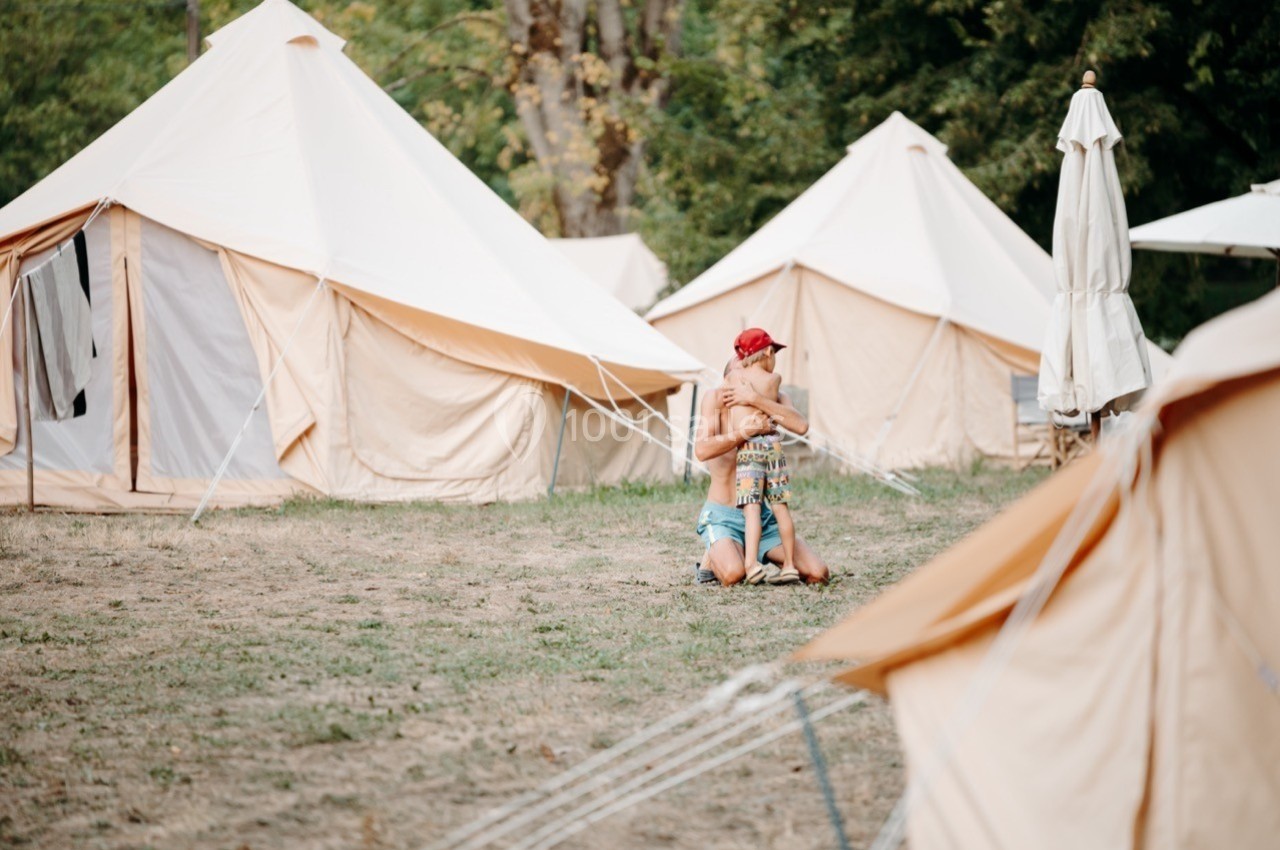 Personne assise sur l'herbe devant des tentes beige, entourée d'arbres en arrière-plan.