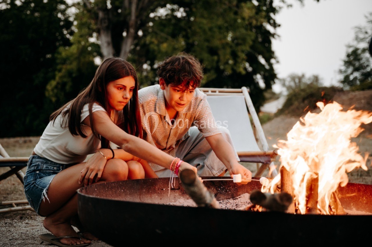 Deux adolescents assis près d'un brasero allument un feu avec des branches, en plein air, entourés de verdure.