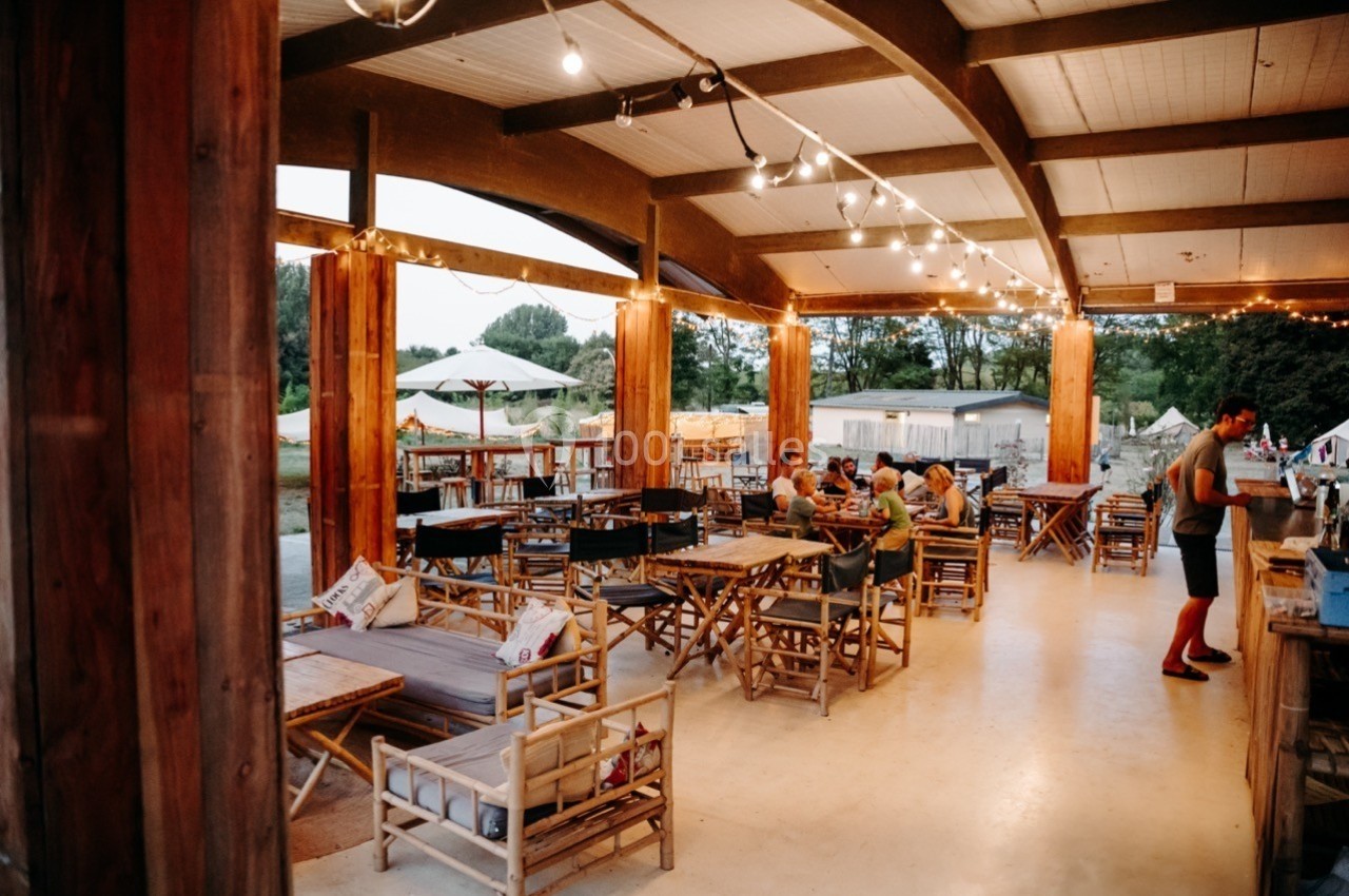 Terrasse couverte avec tables et chaises en bois, éclairée par des guirlandes lumineuses, quelques personnes assises.