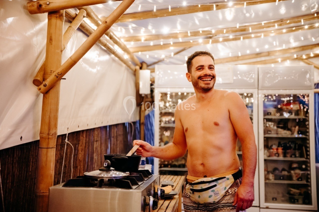 Un homme torse nu cuisine sur une plaque de cuisson sous un abri en bois décoré de guirlandes lumineuses.