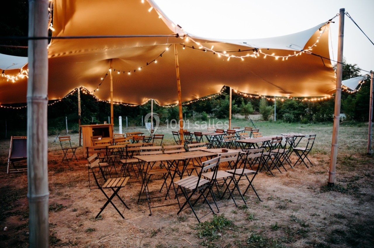 Chaises et tables en bois disposées sous une grande tente éclairée par des guirlandes lumineuses, en extérieur.