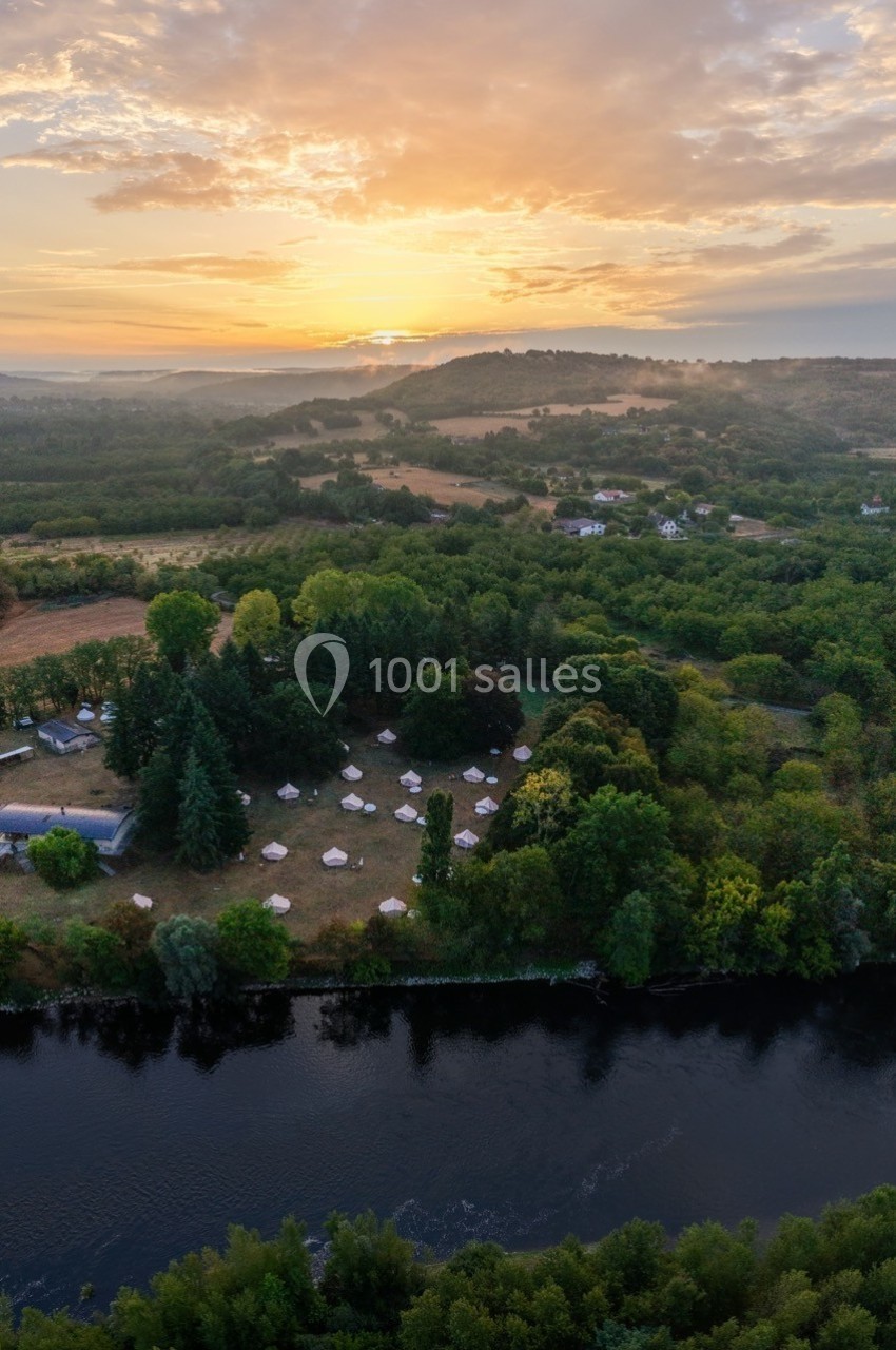 Vue aérienne d'un paysage rural au lever du soleil avec une rivière, des arbres et des tentes blanches dispersées.