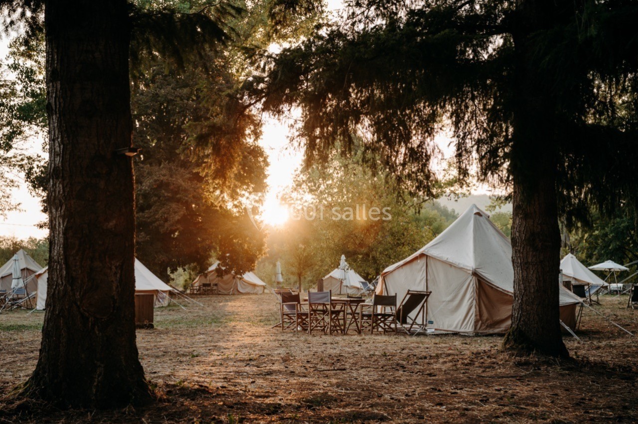 Tentes en toile installées dans une clairière boisée au coucher du soleil, avec des chaises disposées au centre.