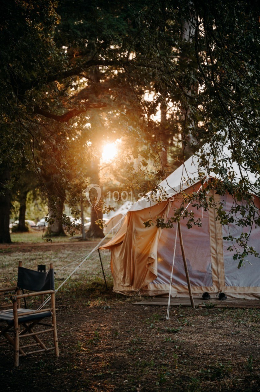 Tente beige installée sous des arbres dans un cadre naturel au coucher du soleil, avec une chaise pliable au premier plan.
